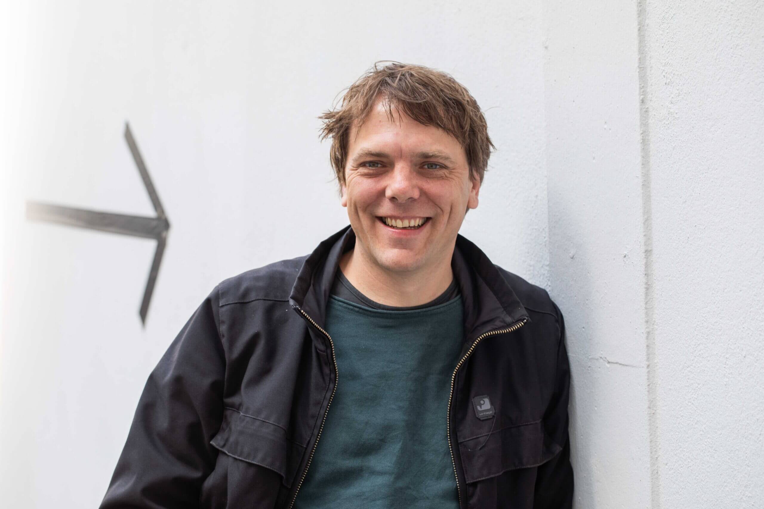 Mann in schwarzer Jacke lächelt vor einer weißen Wand mit Pfeil.